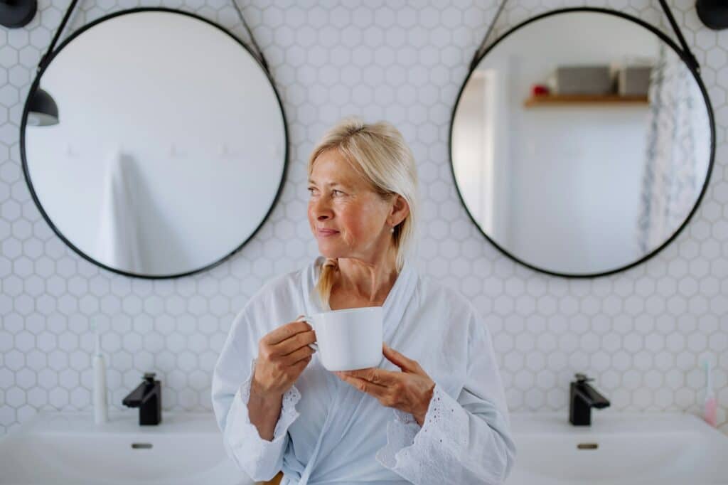 Beautiful senior woman in bathrobe drinking tea in bathroom, relax and wellness concept.